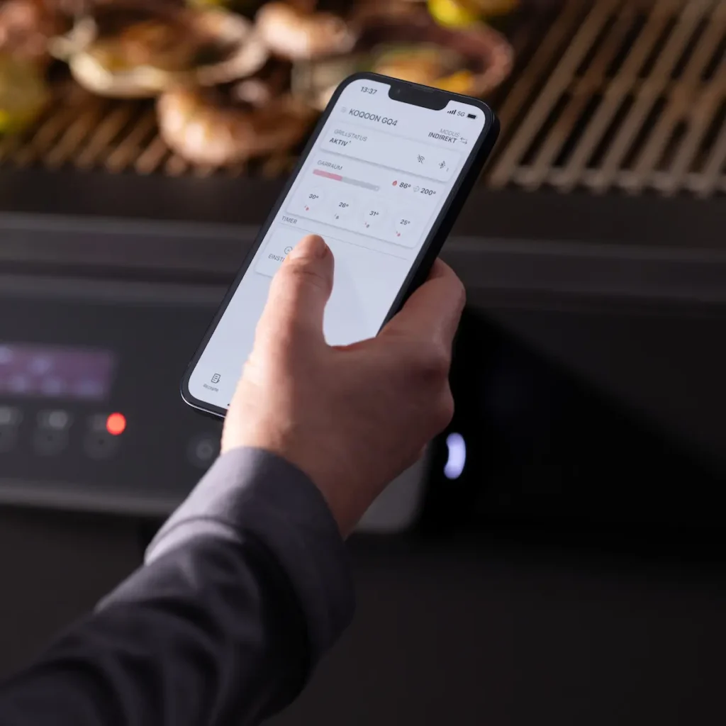 Hand hält ein Smartphone mit der KOQOON GQ4 App zur Steuerung des Grills.